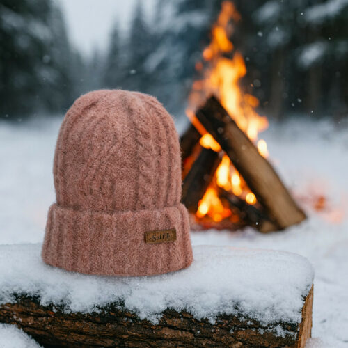 er staat een rode muts op een onder gesneeuwde boomstam en daar achter staat een kampvuurtje te branden