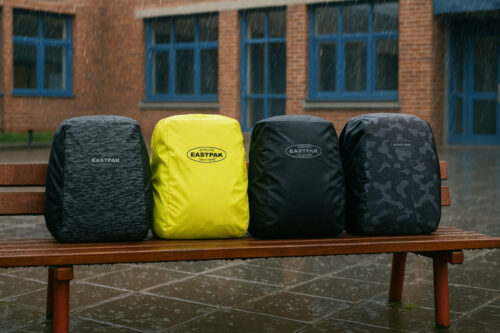 je ziet een bankje voor een school met daar op vier tassen en om die tassen zitten regenhoezen van eastpak