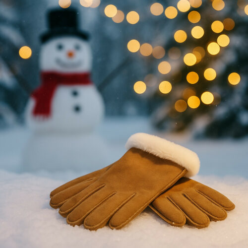 dit zijn cognac kleurende handschoenen die liggen in de sneeuw met als achtergrond een sneeuwpop die heeft een rode sjaal om en een zwarte hoed op en daarnaast nog een leuke boom met lichtjes erin