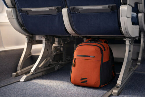 je ziet een Samsonite Moderny Underseater Backpack XS in de kleur rood die onder een stoel van een vliegtuig staat
